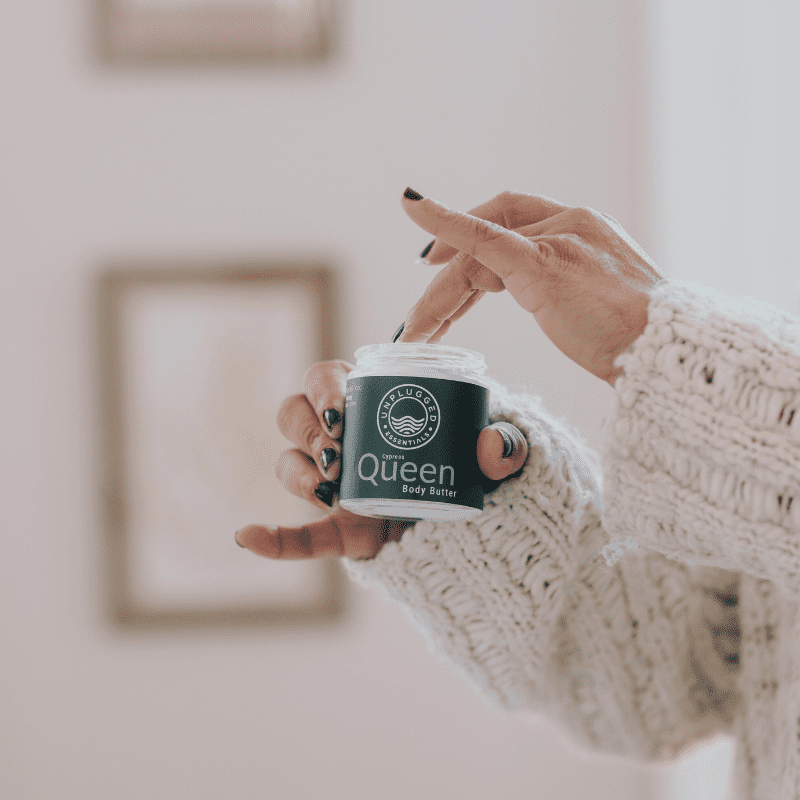 A woman dipping her fingers into the Queen Body Butter by Unplugged Essentials about to apply it to her body.